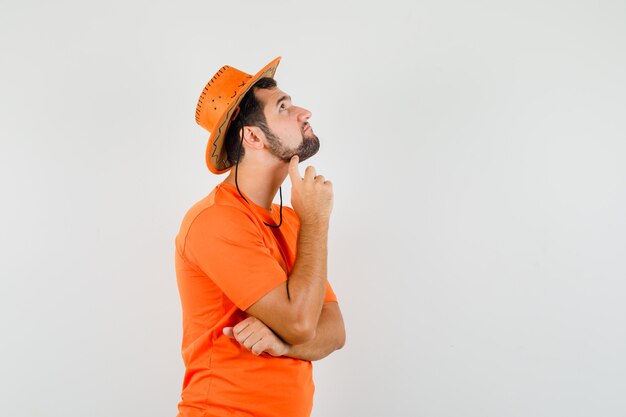 Jovem olhando em t-shirt laranja, chapéu e olhando pensativo, vista frontal.