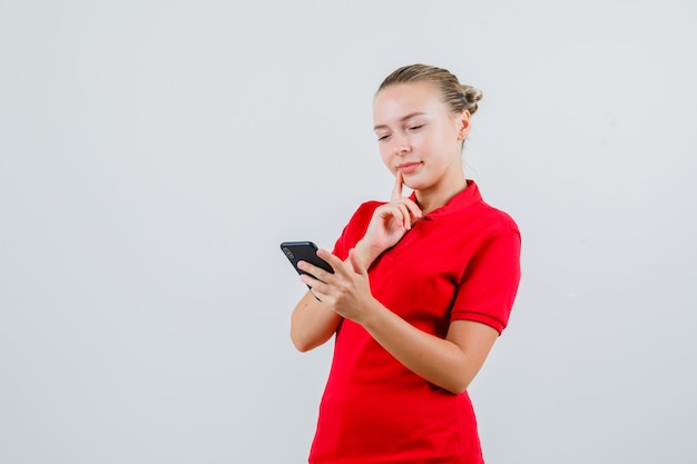 Jovem olhando para o celular com uma camiseta vermelha e parecendo pensativa