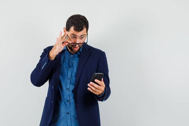 Jovem olhando para o celular por cima dos óculos na camisa, jaqueta e parecendo chocado, vista frontal.