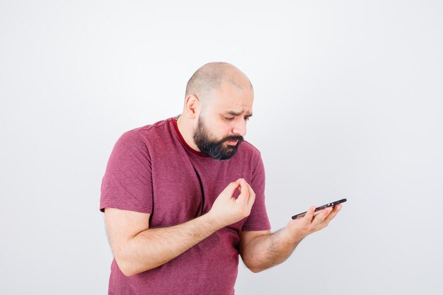 Jovem olhando para o telefone e esticando a mão, explicando algo para alguém em uma camiseta rosa e parecendo triste, vista frontal.