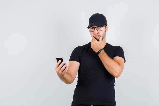 Jovem olhando para um smartphone com a mão no queixo em uma camiseta preta