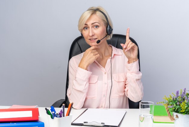 Jovem operadora de call center impressionada usando fone de ouvido, sentada à mesa com ferramentas de escritório apontando para cima