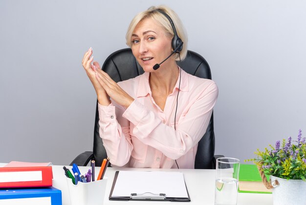 Jovem operadora de call center satisfeita usando fone de ouvido, sentada à mesa com ferramentas de escritório de mãos dadas isoladas na parede branca