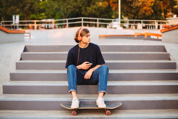 Jovem patinadora em fones de ouvido laranja com celular nas mãos, sentado nas escadas no moderno parque de skate