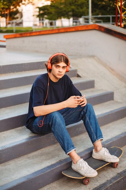 Jovem patinadora em fones de ouvido laranja segurando o celular nas mãos olhando pensativamente na câmera sentado na escada com skate no parque de skate