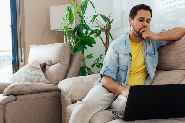 Jovem pensando em algo em casa em seu sofá e olhando para um laptop