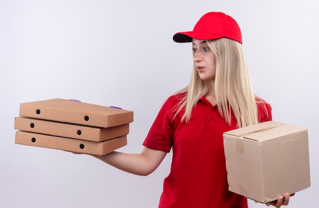 Jovem pensando em entrega, vestindo camiseta vermelha e boné, segurando uma caixa, olhando para a caixa de pizza em sua mão na parede branca isolada