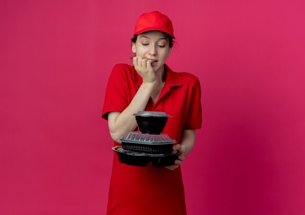 Foto grátis jovem pensativa, bonita, entregadora de uniforme vermelho e boné, segurando e olhando para recipientes de comida e dedos mordendo isolados no fundo carmesim com espaço de cópia