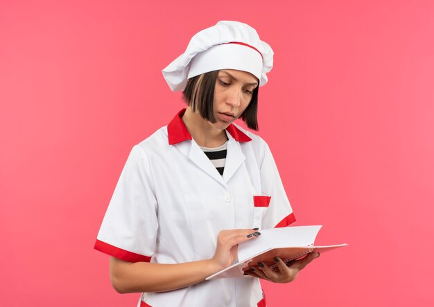 Jovem pensativa cozinheira em uniforme de chef segurando e olhando para o bloco de notas isolado na parede rosa