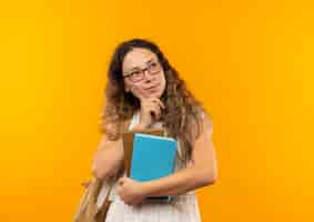 Foto grátis jovem pensativa e bonita colegial usando óculos e bolsa nas costas segurando livros, colocando a mão no queixo, olhando para o lado isolado na parede amarela