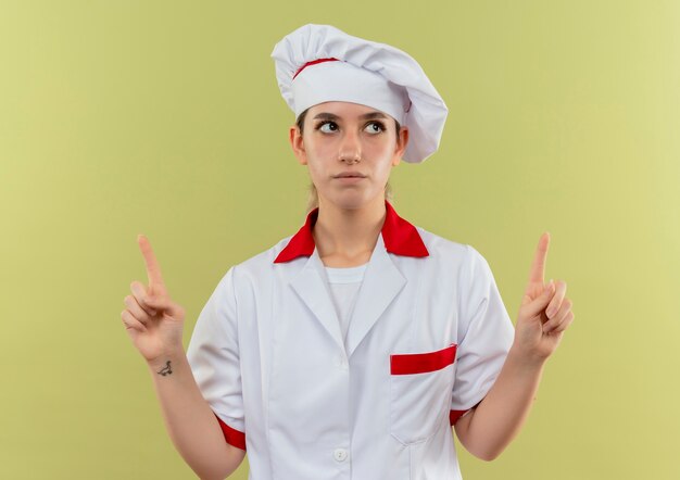 Jovem pensativa e bonita cozinheira em uniforme de chef apontando e olhando para cima, isolada no espaço verde