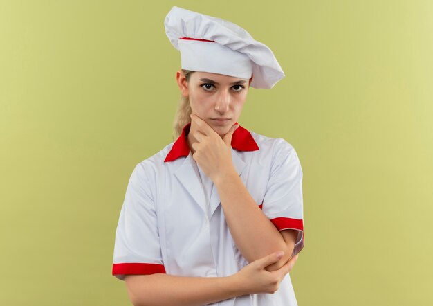 Jovem pensativa e bonita cozinheira em uniforme de chef, colocando a mão no queixo, parecendo isolada em um espaço verde