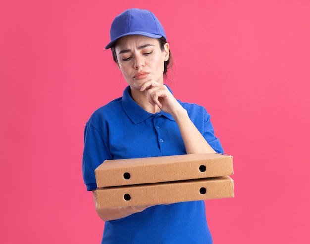 Jovem pensativa entregadora de uniforme e boné segurando e olhando para pacotes de pizza tocando o queixo isolado na parede rosa com espaço de cópia