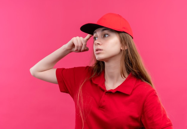 Jovem pensativa, entregadora de uniforme vermelho, colocando o dedo na cabeça, olhando para o lado esquerdo na parede rosa