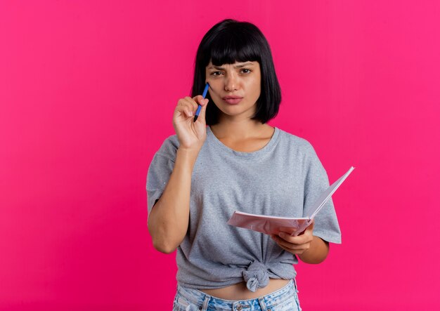 Jovem pensativa, morena, caucasiana, coloca uma caneta no rosto e segura um caderno olhando para a câmera isolada no fundo rosa com espaço de cópia