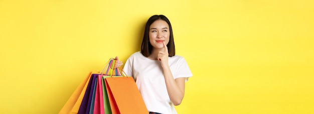 Jovem pensativa sorrindo intrigada mostrando sacolas de compras pensando em comprar algo em pé