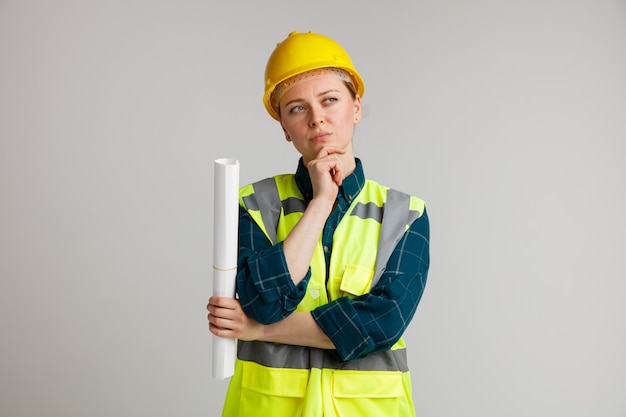 Jovem pensativa, trabalhadora da construção civil, usando capacete e colete de segurança, segurando um papel, mantendo a mão no queixo, olhando para o lado