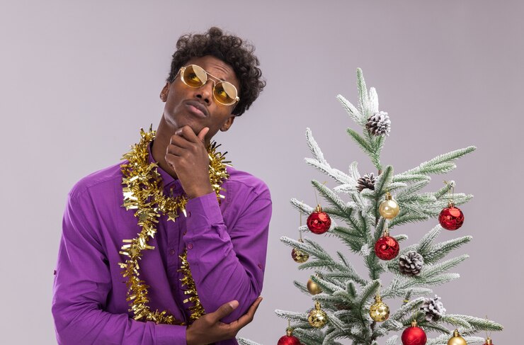 Foto grátis jovem pensativo, afro-americano, usando óculos com guirlanda de ouropel no pescoço, em pé perto da árvore de natal decorada, segurando o queixo, olhando para cima, isolado no fundo branco