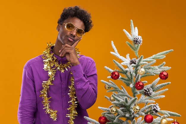 Jovem pensativo, afro-americano, usando óculos com uma guirlanda de ouropel em volta do pescoço, em pé perto de uma árvore de natal decorada em fundo laranja