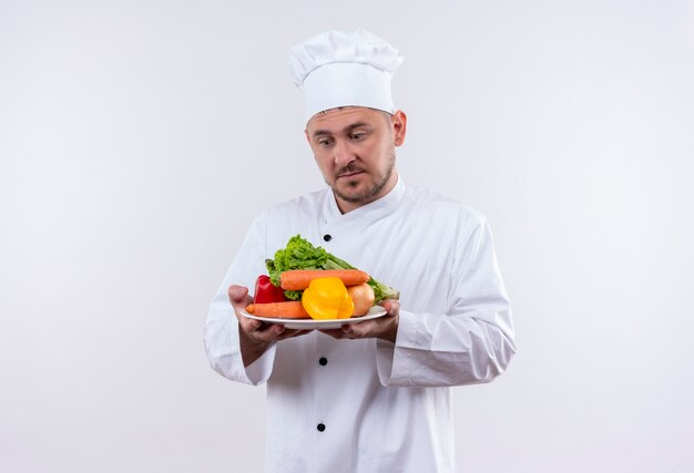 Jovem pensativo bonito cozinheiro em uniforme de chef segurando o prato com legumes olhando para eles no espaço em branco isolado