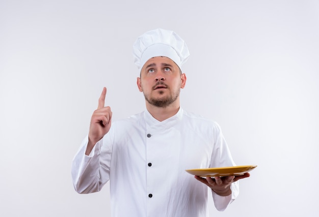 Jovem pensativo bonito cozinheiro em uniforme de chef segurando o prato, olhando e apontando para cima no espaço em branco isolado