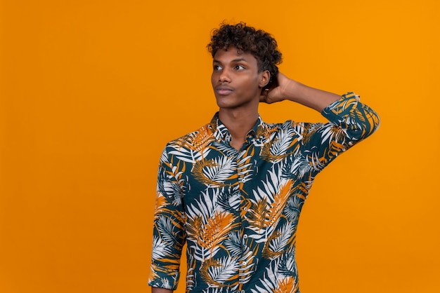 Jovem pensativo, bonito, de pele escura, com cabelo encaracolado e camisa estampada de folhas, mantendo as mãos na cabeça pensando em um fundo laranja