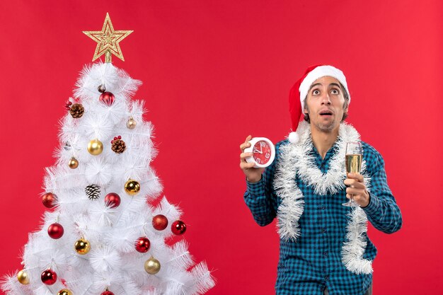 Jovem pensativo com chapéu de Papai Noel, levantando uma taça de vinho e segurando o relógio em pé perto da árvore de Natal no vermelho