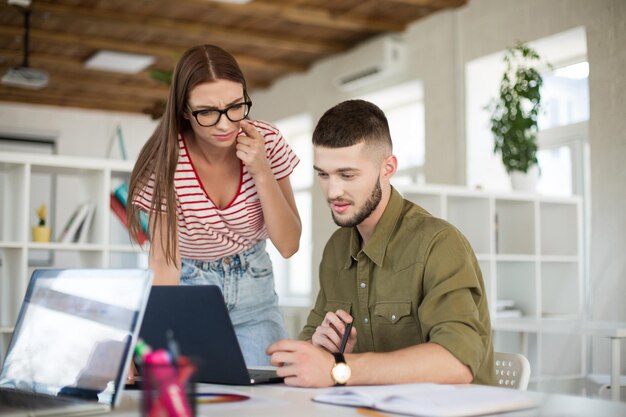 Jovem pensativo de camisa e mulher de camiseta listrada e óculos trabalhando sonhadoramente em conjunto com o laptop Empresários criativos passando tempo no trabalho no escritório moderno e aconchegante