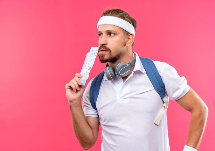 Foto grátis jovem pensativo e esportivo bonito usando bandana, pulseira e bolsa traseira com fones de ouvido no pescoço, segurando passagens aéreas e olhando para o lado isolado no espaço rosa
