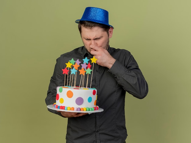 jovem pensativo festeiro vestindo camisa preta e chapéu azul segurando e olhando para um bolo agarrado no nariz isolado no verde oliva