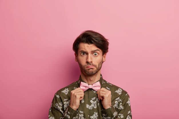 Jovem perplexo ergue as sobrancelhas, tem cerdas, ajusta gravata borboleta, veste camisa da moda com estampa moderna, se veste com bom gosto, fica contra o espaço rosado. Pessoas