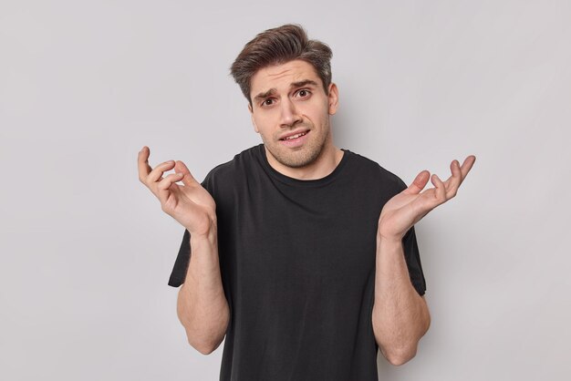 Jovem perplexo inseguro espalha as palmas das mãos sente-se incomodado ao fazer a escolha parece sem noção usa camiseta preta casual isolada sobre fundo branco. Modelo masculino questionado fica dentro de casa