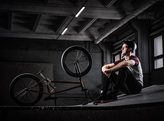 Jovem piloto de Bmx relaxando depois de praticar truques com sua bicicleta em um skatepark dentro de casa