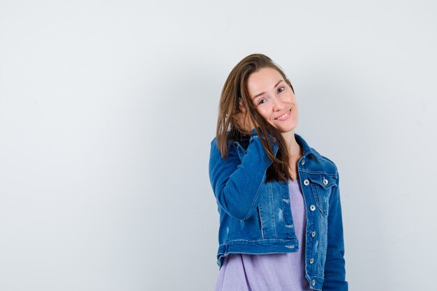 Jovem posando com a mão na cabeça em t-shirt, jaqueta e olhando graciosa, vista frontal.