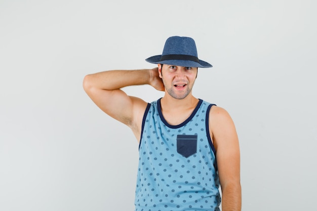 Jovem posando com a mão no pescoço em camiseta azul, chapéu e parecendo alegre. vista frontal.