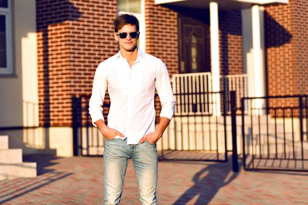 Jovem posando na rua, look casual inteligente, cores quentes em tons ensolarados, jovem empresário andando sozinho, óculos escuros da moda, camisa branca.