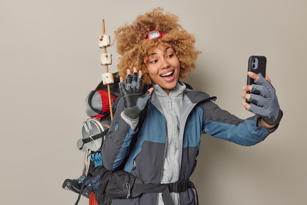 Jovem positiva usa jaqueta de farol e luvas acena olá tem videoconferência com amigo carrega mochila retorna de acampamento isolado sobre fundo cinza pessoas desejo de viajar