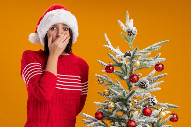 Jovem preocupada com chapéu de papai noel em pé perto da árvore de natal decorada, segurando a mão na boca de outra atrás das costas, olhando para a câmera isolada em fundo laranja