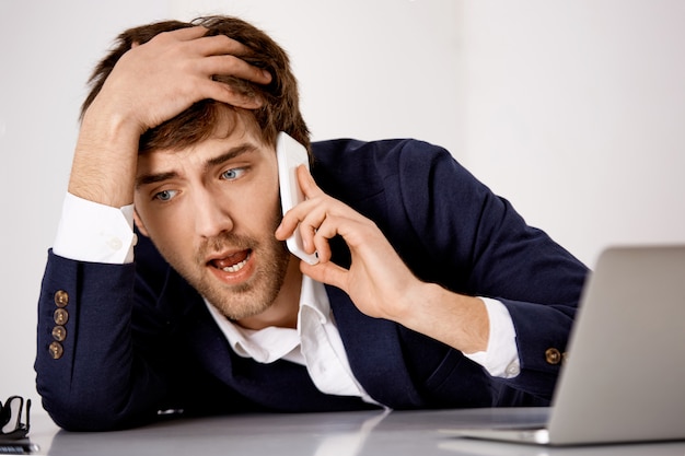 Jovem problemático, incline-se na mesa angustiada, tendo uma conversa séria no telefone, grande problema com o parceiro de negócios