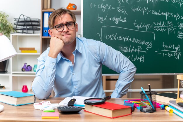 Jovem professor de óculos, confuso e muito ansioso, sentado na mesa da escola com livros e anotações na frente do quadro-negro na sala de aula
