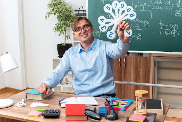 Jovem professor de óculos mostrando matrículas explicando a aula, sorrindo, sentado na mesa da escola com livros e anotações na frente do quadro-negro na sala de aula