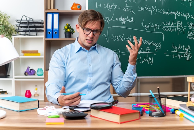 Jovem professor de óculos, parecendo confuso e desapontado, sentado na mesa da escola com livros e anotações na frente do quadro-negro na sala de aula