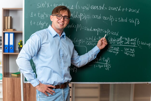 Jovem professor de óculos segurando giz, explicando a lição em pé perto do quadro-negro com fórmulas matemáticas na sala de aula