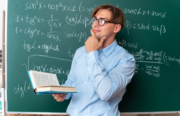 Jovem professor de óculos segurando um livro, olhando para o lado com a mão no queixo, pensando em pé perto do quadro-negro com fórmulas matemáticas em sala de aula