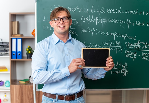 Jovem professor de óculos segurando uma pequena lousa e sorrindo confiante em pé perto da lousa com fórmulas matemáticas