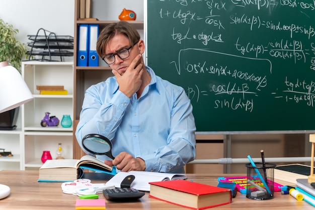 Jovem professor de óculos sentado na mesa da escola com livros e notas olhando através de uma lupa para um livro com uma cara séria na frente do quadro-negro na sala de aula