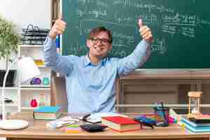 Foto grátis jovem professor de óculos, sorrindo alegremente, mostrando os polegares para cima, sentado na mesa da escola com livros e anotações na frente do quadro-negro na sala de aula