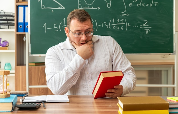 jovem professor preocupado usando óculos, sentado à mesa com material escolar na sala de aula, segurando e olhando para o livro fechado, segurando a mão no queixo