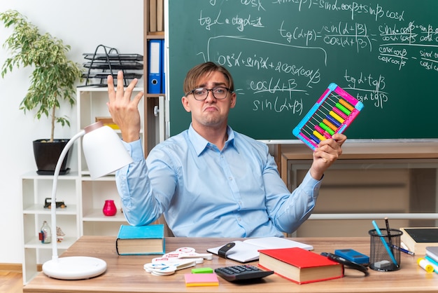 Foto grátis jovem professor usando óculos e contas, parecendo confuso, preparando a aula, sentado na mesa da escola com livros e anotações na frente do quadro-negro na sala de aula