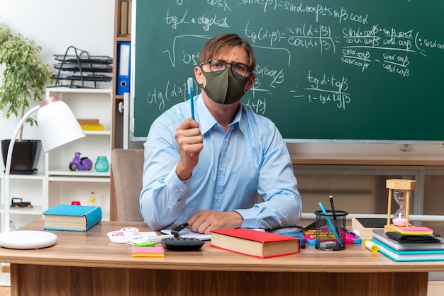 Jovem professor usando óculos e máscara protetora facial mostrando um lápis com cara séria, sentado na mesa da escola com livros e anotações na frente do quadro-negro na sala de aula